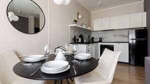 Un comedor moderno con una mesa redonda preparada para cuatro, sillas blancas y un mantel negro. Se ve una cocina con armarios blancos y un refrigerador negro al fondo.