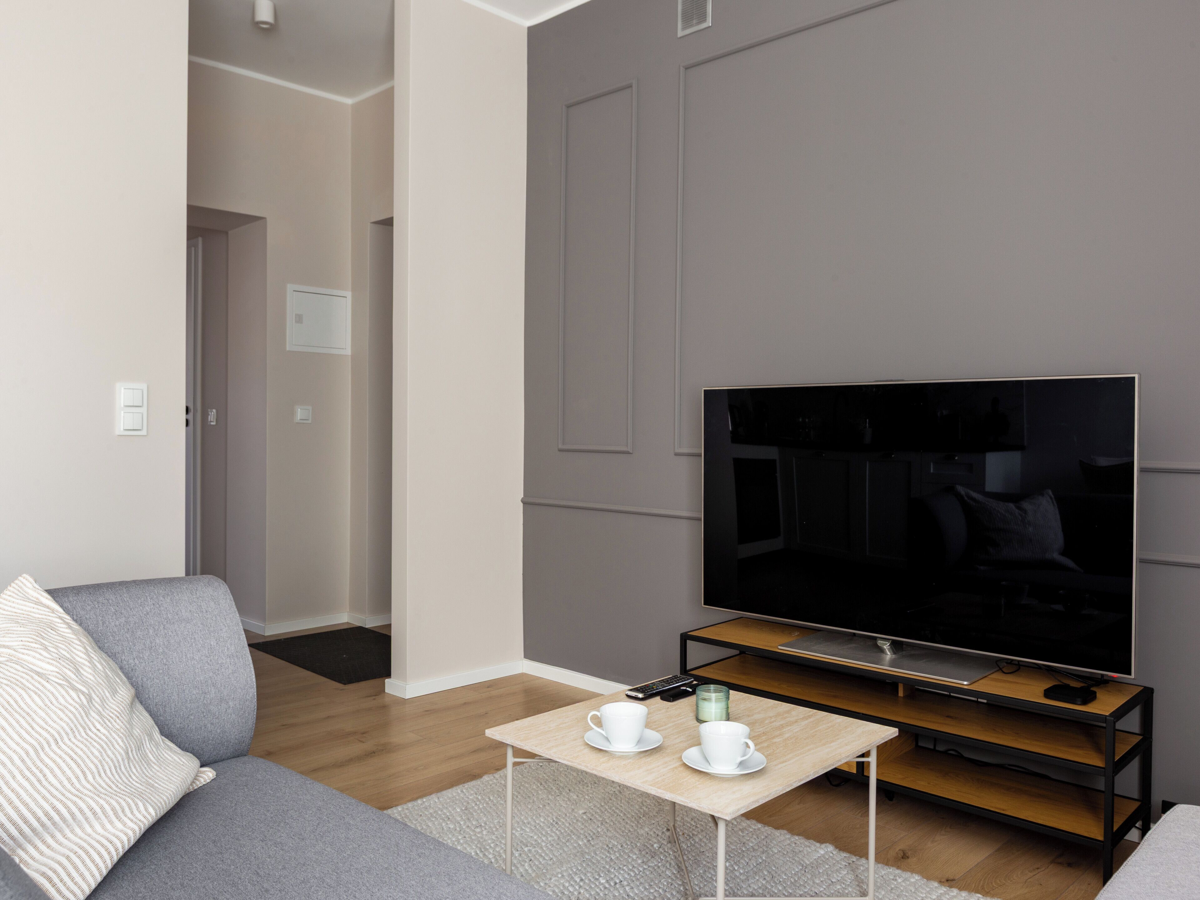 A modern living room with a grey sofa, a coffee table with cups, a TV on a wooden stand, and a door leading to another room.