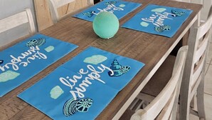 Ein Esstisch mit vier blauen Platzsets mit nautischen Motiven und der Aufschrift 'Live Simply'. Auf dem Tisch liegt ein grüner Ball.