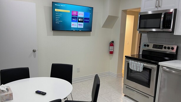 A modern kitchen with a flat-screen TV displaying a Roku TV interface, a stainless steel microwave, an oven, and a dining area with a table and chairs.