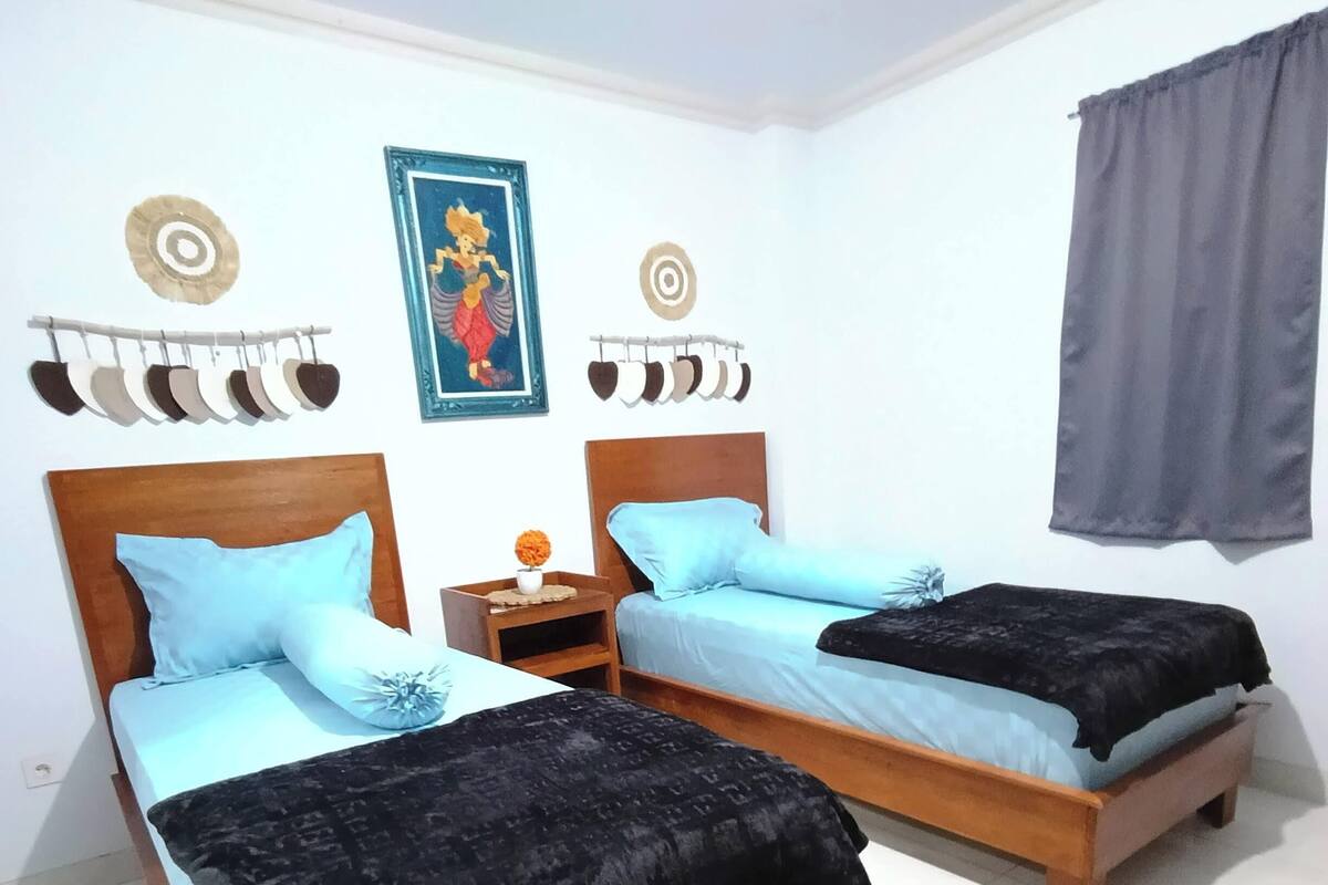 Een kamer met twee houten bedden, elk voorzien van een blauwe dekbedovertrek en zwarte kussens. Er staat een klein nachtkastje met een decoratief object erop. De muren zijn wit en er hangen twee ingelijste foto's boven de bedden.