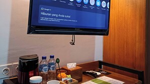 Een hotelkamer met een flatscreen-tv aan de muur, waarop diverse streamingapps getoond worden. Er staat een houten tafeltje met een koffiezetapparaat, twee waterflessen, een beker en een pak zakdoekjes.
