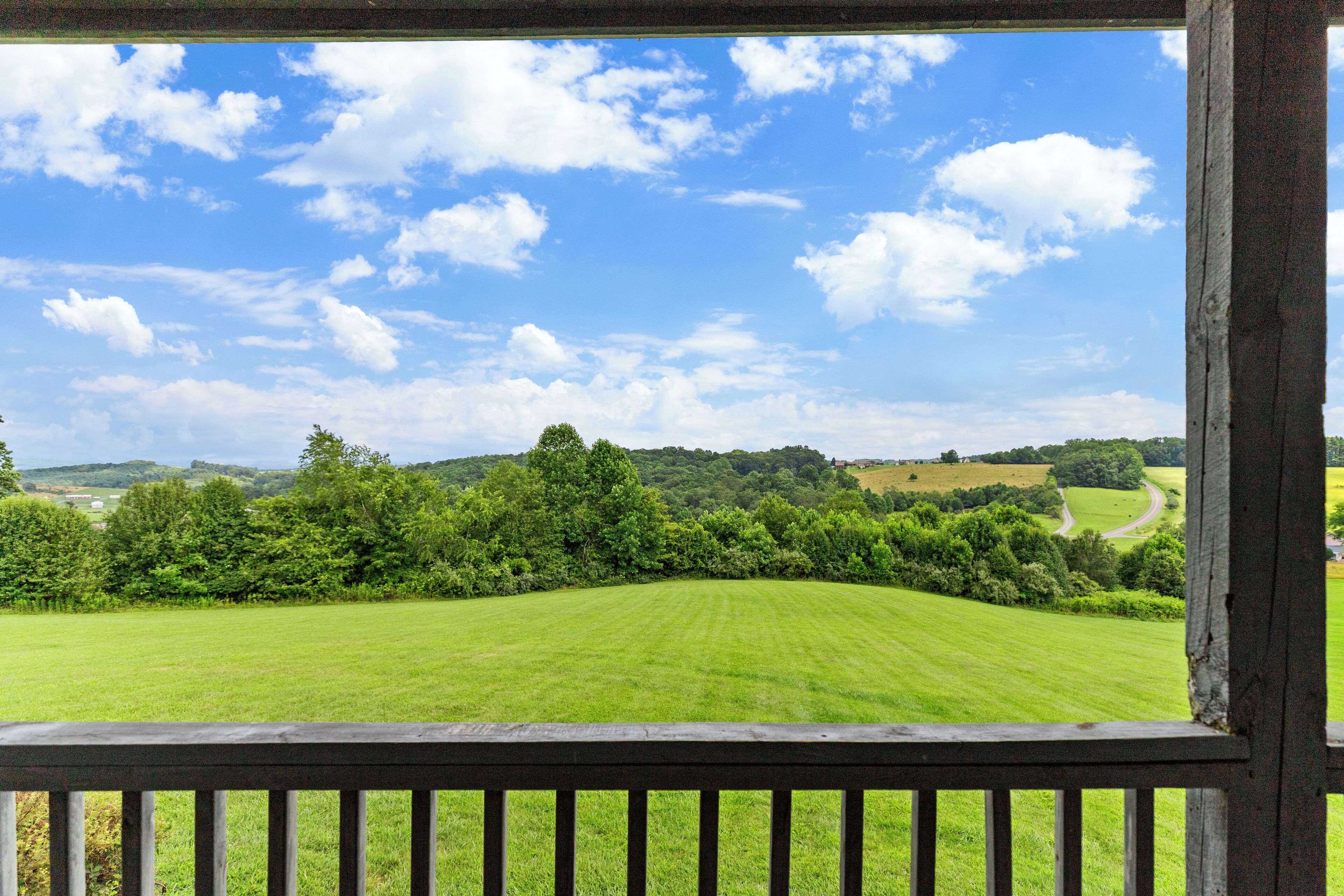 A view from a wooden balcony overlooking a green field and a winding road.