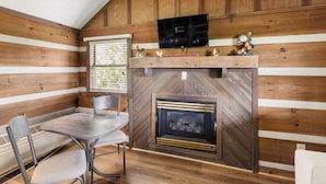 Interior acogedor de una cabaña con chimenea, un televisor montado encima y una zona de comedor con mesa y sillas.