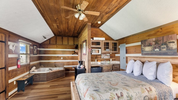 Un dormitorio de estilo cabaña de madera con una cama grande, un ventilador de techo de madera, una bañera integrada y una pequeña cocina.