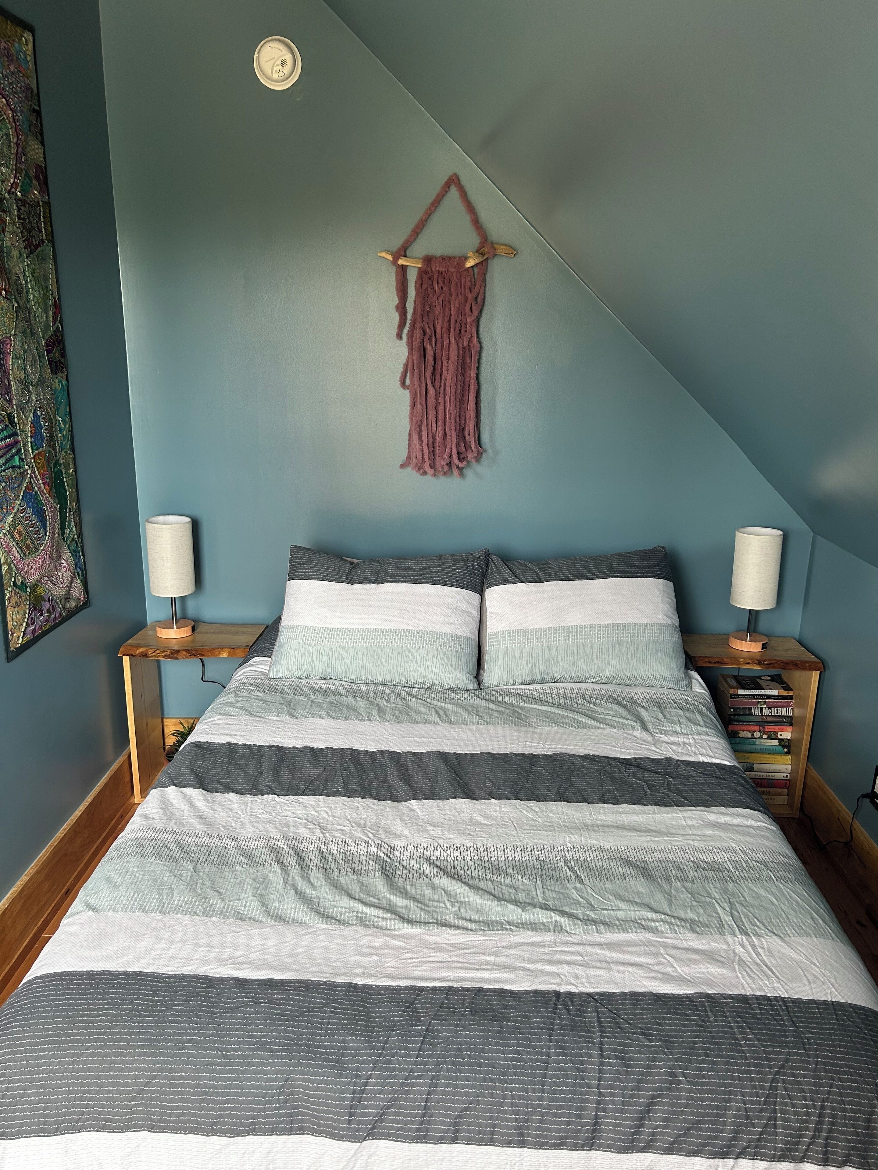 Ein Schlafzimmer mit einem Bett, zwei Nachttischen, einer Wanddekoration aus Makramee und einem Bücherregal.
