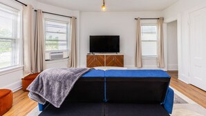 Una habitación con una cama, un televisor sobre un mueble de madera y ventanas grandes con cortinas.