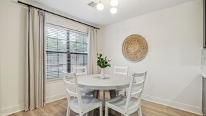 Een eetgedeelte met een ronde tafel, witte stoelen, een potplant en een wanddecoratie van geweven materiaal.
