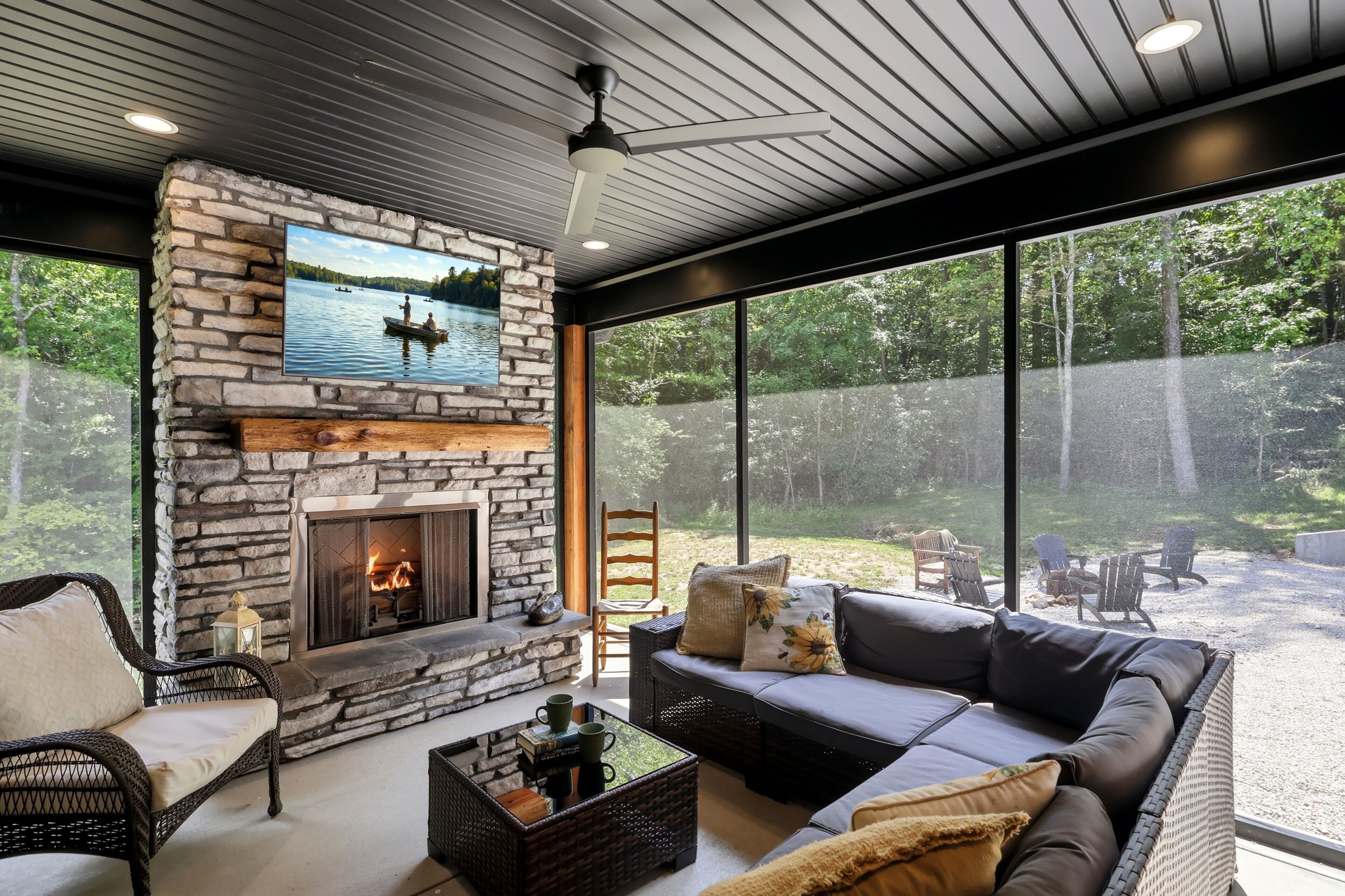 A modern living room with a stone fireplace, a large window overlooking a forest, and a ceiling fan.