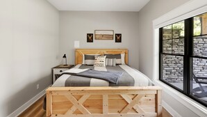 A bedroom with a wooden bed frame, a nightstand, a lamp, a ceiling fan, and a large window with a stone wall view. - Private Nolin Lake Home with FirePit, Ice Maker, Arcade & Electronic Game Table. (Clarkson)