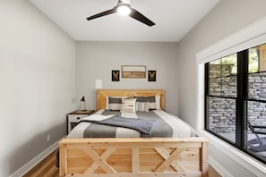 Une chambre à coucher avec un cadre de lit en bois, une table de chevet, une lampe, un ventilateur de plafond et une grande fenêtre donnant sur un mur en pierre.