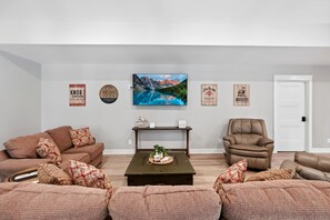 A living room with a brown leather sofa, a green coffee table, and a flat-screen TV mounted on the wall. - Private Nolin Lake Home with FirePit, Ice Maker, Arcade & Electronic Game Table. (Clarkson)