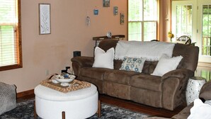 Amplio salón con techo alto, escalera de madera y una zona de estar acogedora con un sofá y reposapiés marrones.