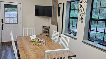 A dining area with a wooden table, white chairs, a flat-screen TV mounted on the wall, and windows with views of greenery.