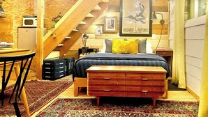 A bedroom with a sloped ceiling, a bed with a dark blue bedspread, a wooden bench, a desk with a chair, and a small table. - Tucked Away Stay in East ATX (Austin)