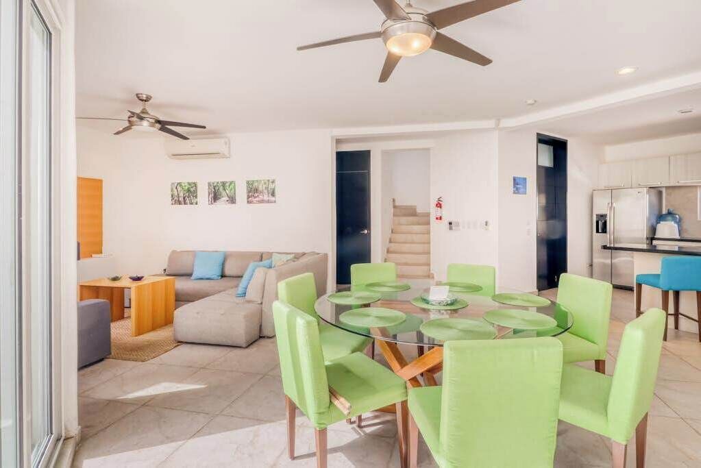A modern living room with a glass-top dining table, green chairs, a sofa, and a ceiling fan.