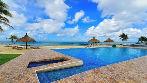 A pool area with a clear view of the ocean, stone flooring, and thatched umbrellas.