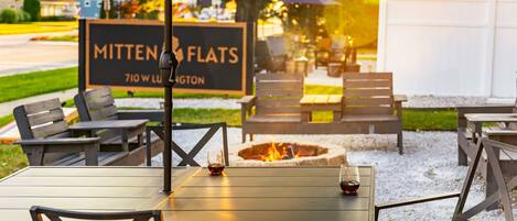 An outdoor seating area with tables and chairs, a fire pit, and a sign reading "Mitten Flats 710 W. LUGTON".