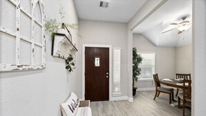 Um quarto com porta de madeira, um banco com almofada de 'Welcome', uma mesa de jantar com cadeiras e um ventilador de teto.