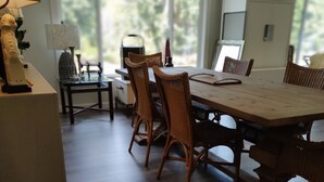A dining area with a wooden table and wicker chairs, a lamp, and a view of greenery outside.