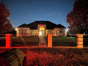 Een groot, goed verlicht gebouw met een hekpoort, omringd door een aangelegde tuin en bomen met herfstkleur.