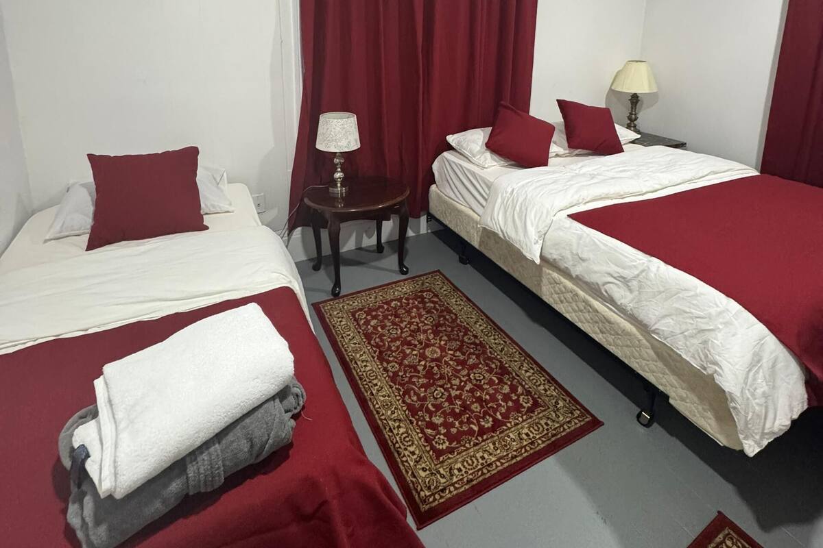 Ein Hotelzimmer mit zwei Betten, roten Vorhängen, einem kleinen Tisch mit Lampe und einem gemusterten Teppich.