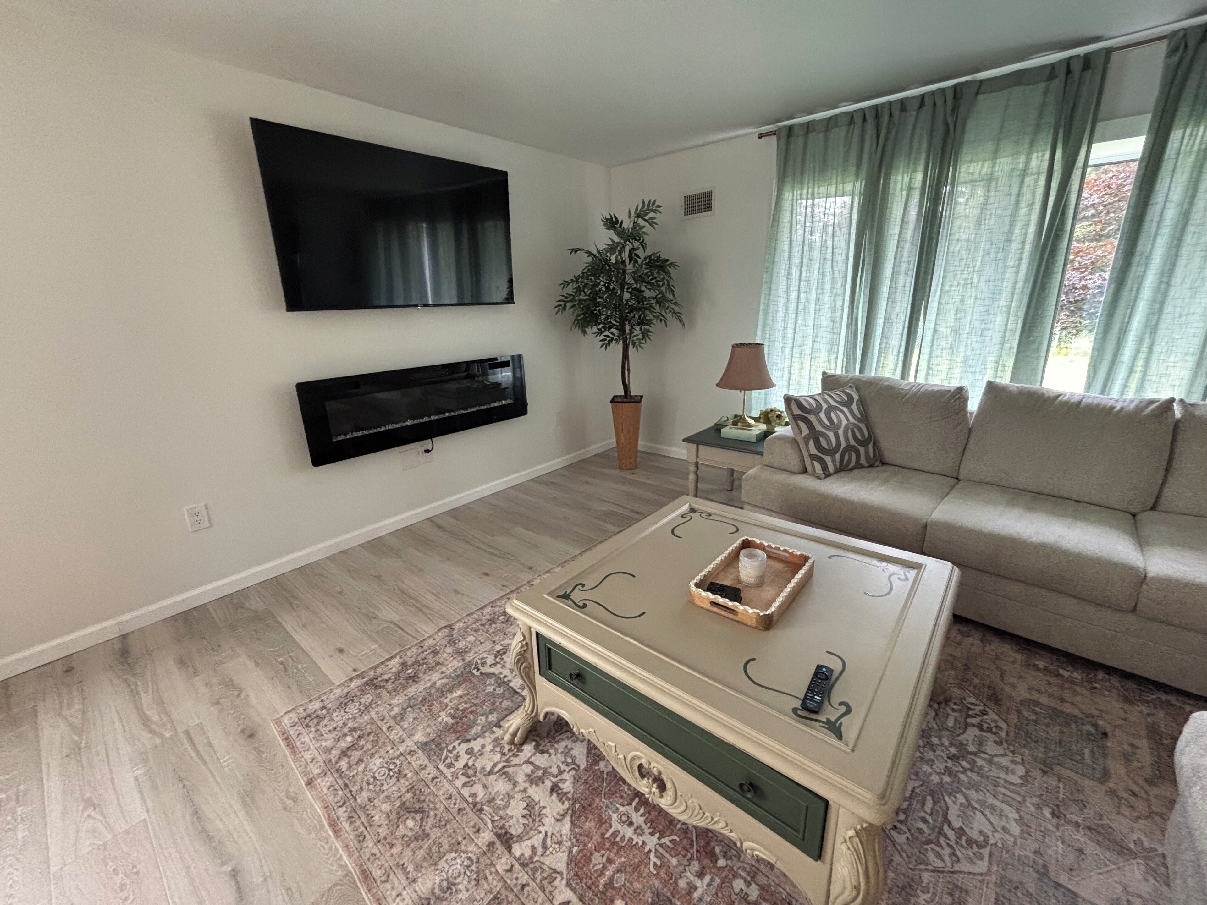 A living room with a flat-screen TV mounted on the wall, a sofa, a coffee table with a tray, a potted plant, and a lamp on a side table.