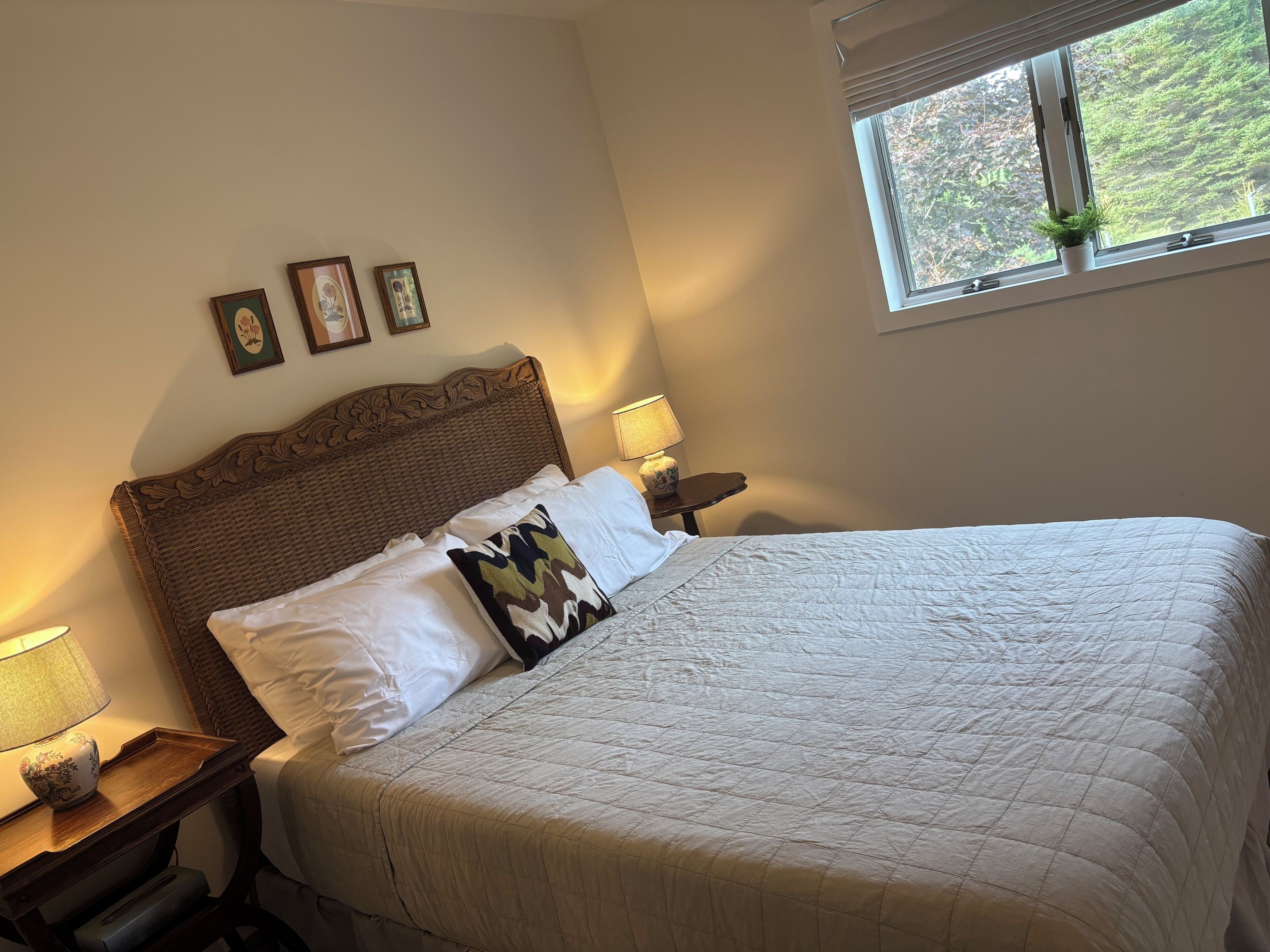Een net opgemaakt bed met een wit geborduurd sprei en twee kussens, een houten nachtkastje met een lamp, en een raam met uitzicht op bomen.