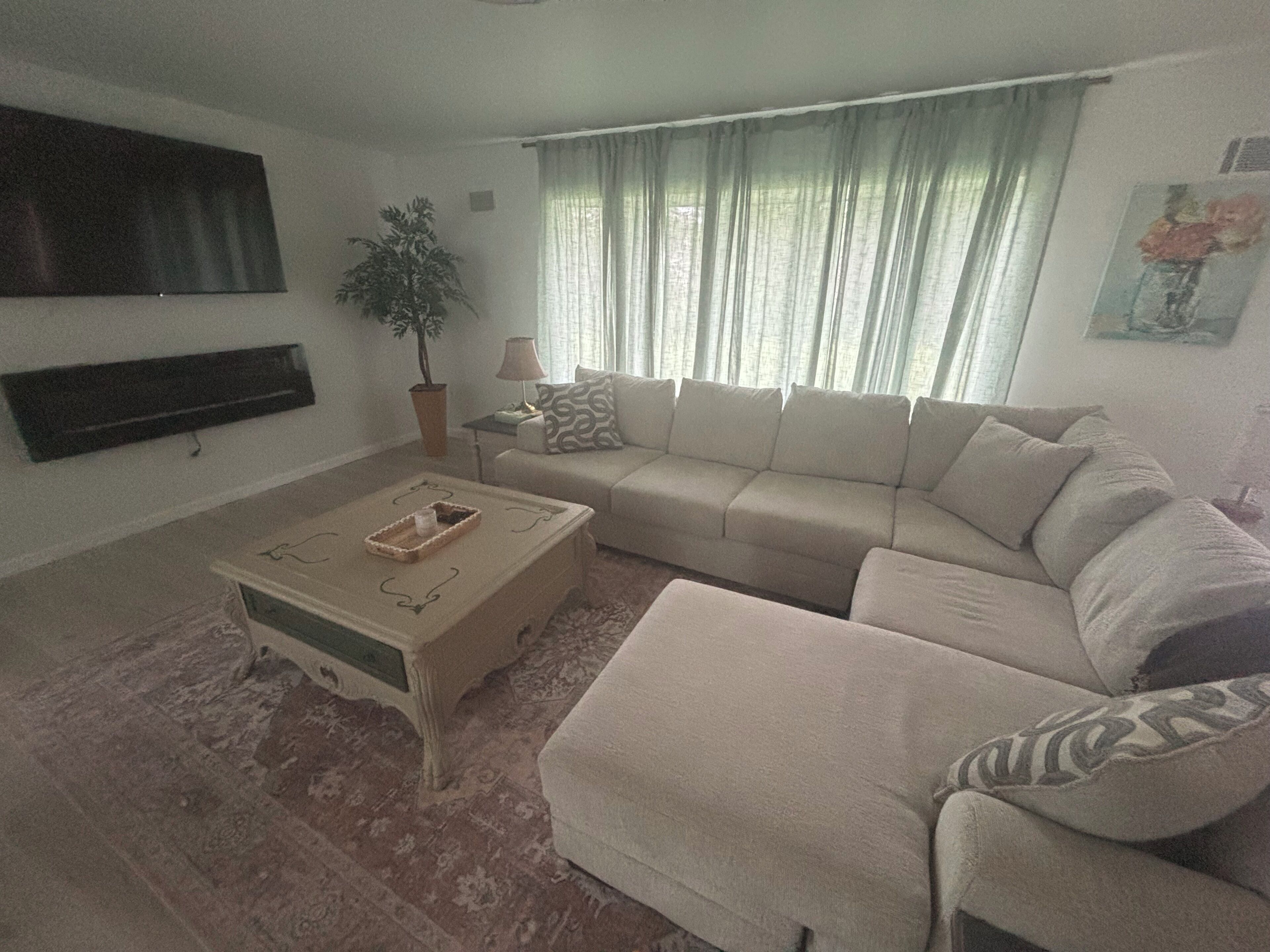 A living room with a sectional sofa, a coffee table, a TV mounted on the wall, a potted plant, and a window with curtains.