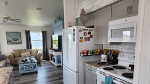 A modern kitchen with white appliances, a dining area with a sofa and coffee table, and a view of the outdoors through a window.