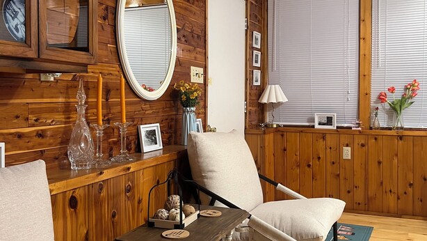 Une pièce chaleureuse avec des lambris en bois, un miroir rond, un fauteuil confortable, une petite table et une fenêtre avec des stores.
