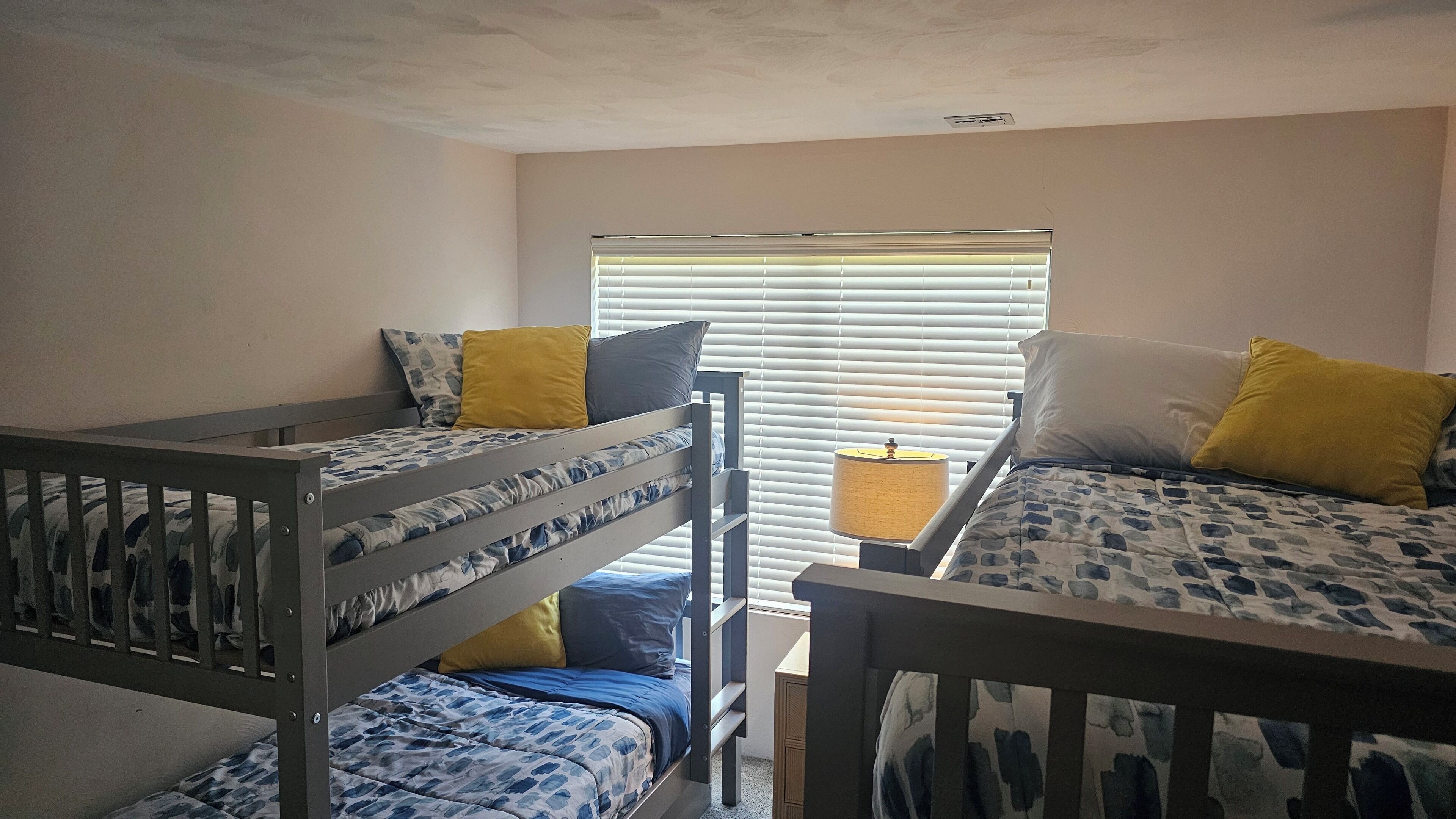 A room with two bunk beds, each with patterned bedding and pillows, a window with blinds, and a bedside lamp.