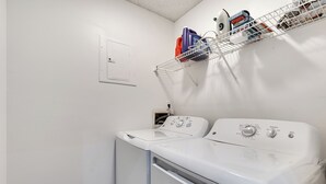 Un área de lavandería con lavadora y secadora de carga frontal, un estante metálico con productos de limpieza y un enchufe en la pared.