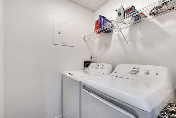 Un área de lavandería con lavadora y secadora de carga frontal, un estante metálico con productos de limpieza y un enchufe en la pared.
