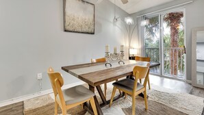 Comedor con mesa de madera, cuatro sillas, ventilador de techo y vista a palmeras a través de amplios ventanales.