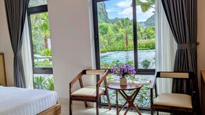 Quarto de hotel com cama, duas cadeiras, uma mesinha com um vaso de flores e amplas janelas com vista para um rio e vegetação.