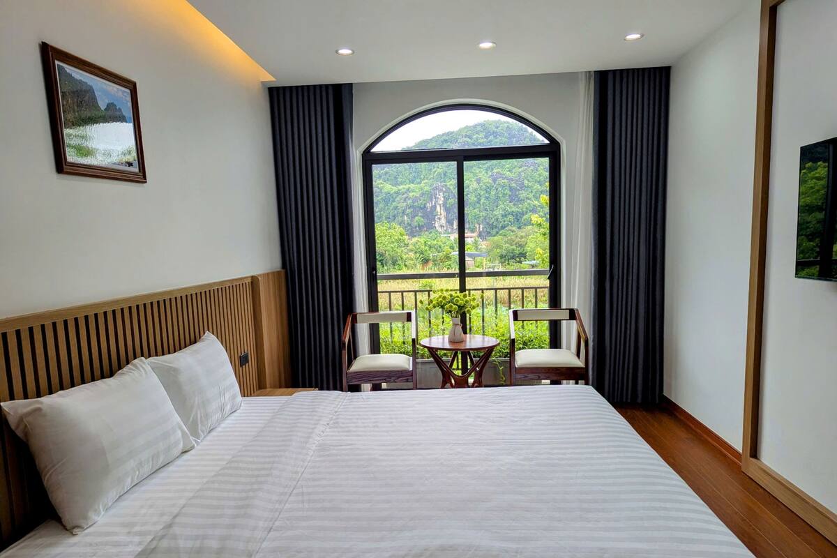 A hotel room with a large bed, a view of greenery through a window, and a small table with chairs.