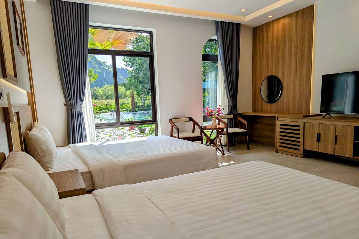 A hotel room with two beds, a desk, a chair, a TV, and large windows with a view of greenery.