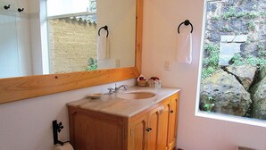 Un baño con tocador de madera, un lavabo, un espejo y una ventana que da a una pared de piedra y follaje.