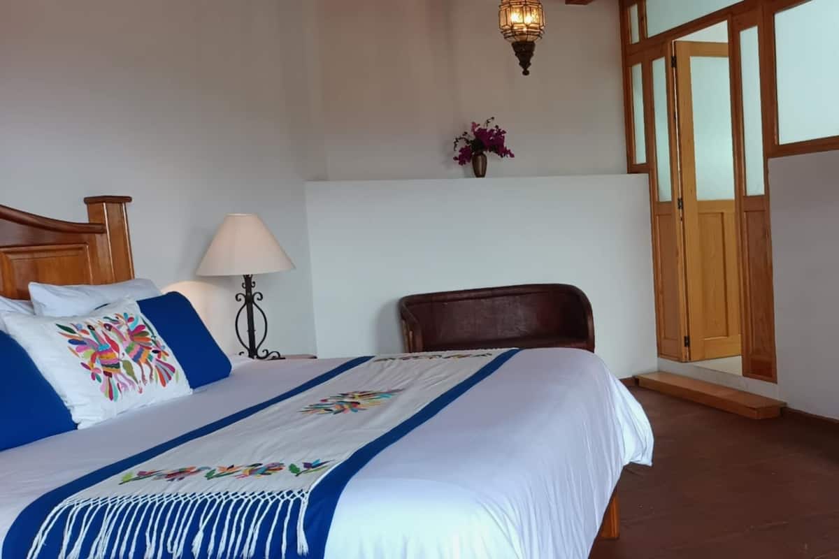 Une chambre à coucher avec un lit, deux lampes de chevet, une commode en bois, une chaise, une porte en bois et un téléviseur fixé au mur.