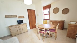 Una habitación con una cama, una mesa de comedor de madera con sillas, un televisor sobre un mueble bajo y una ventana con cortinas.