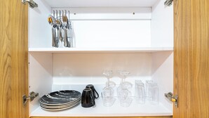 Un armario blanco con estructura de madera, que contiene cubiertos, platos y copas de vino ordenados.