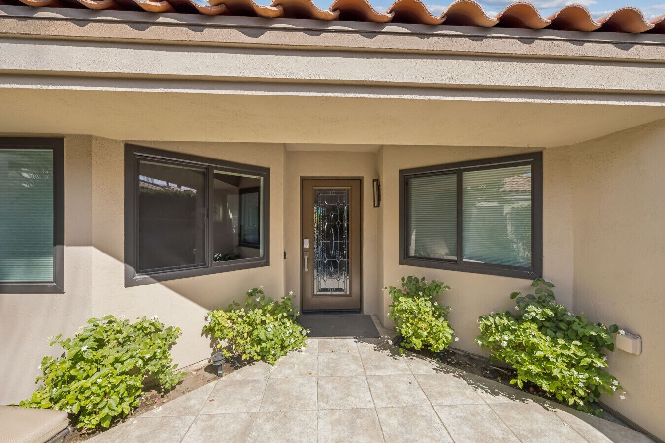 A building entrance with a tiled patio, two windows on either side, and small plants in the entrance area.