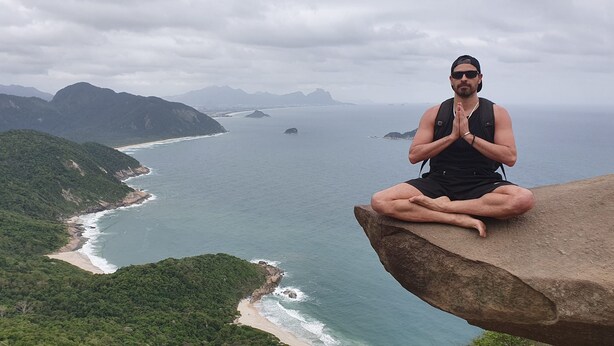 Eine Person meditiert auf einer Klippe mit Blick auf eine Küstenlandschaft, Berge und das Meer.