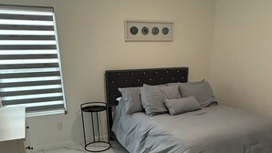 A bedroom with a bed, a bedside table, a lamp, and a window with blinds. -  Brand New, Spacious & Stylish – Your Perfect Getaway Awaits! (Mission)