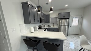 A modern kitchen with grey cabinets, a white countertop, two black bar stools, stainless steel appliances, and a white door with a window. -  Brand New, Spacious & Stylish – Your Perfect Getaway Awaits! (Mission)