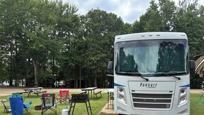 A white coach bus parked in a campsite with picnic tables and camping chairs. - Spacious RV in Brilliant Broken Bow with AC, WiFi, near Day Spa and Winery! (Broken Bow)