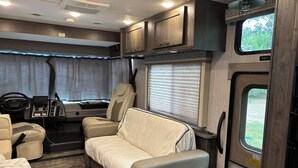 A compact RV interior with a sofa, a small table, a steering wheel, and a window with blinds. - Spacious RV in Brilliant Broken Bow with AC, WiFi, near Day Spa and Winery! (Broken Bow)