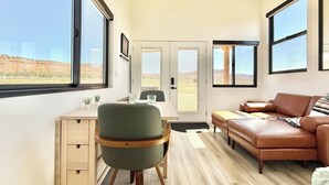 A modern interior with a desk, chair, sofa, and large windows offering a view of the desert landscape. - Journey Pines: Secluded Tiny Home w/ Hot Tub (Kanab)
