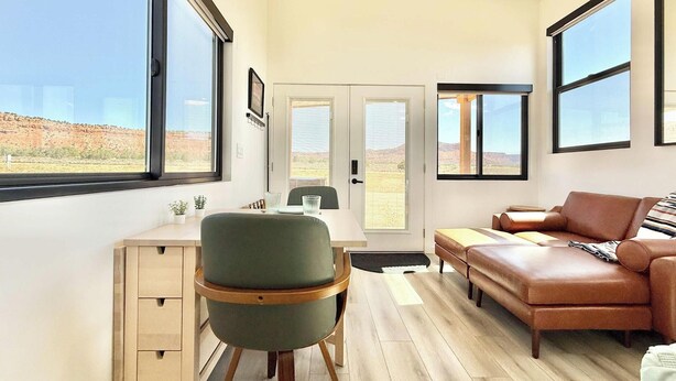 A modern interior with a desk, chair, sofa, and large windows offering a view of the desert landscape. - Journey Pines: Secluded Tiny Home w/ Hot Tub (Kanab)